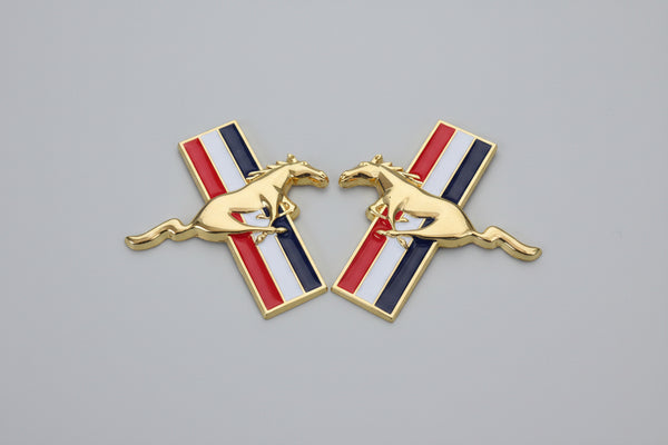 A set of two gold-colored Mustang fender badges with a red, white, and blue stripe and a galloping horse emblem in the center.