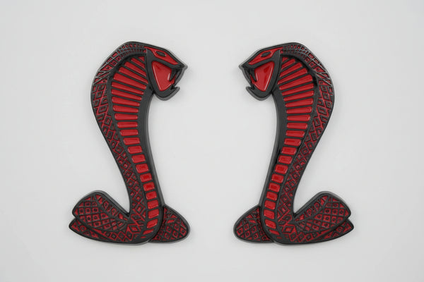 A set of two metal badges in the shape of a cobra, with a black outline and red fill, designed for attachment to a vehicle fender.
